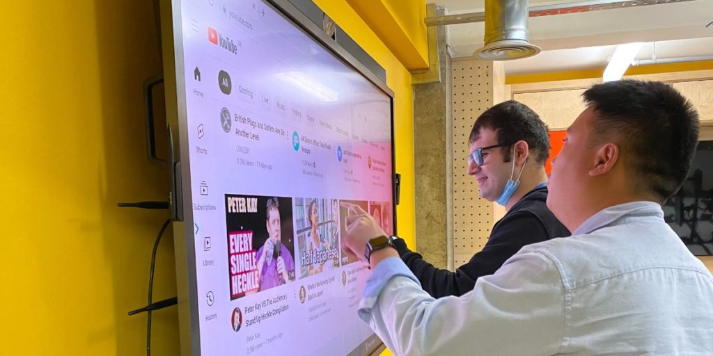 2 young men standing up in front of a big digital screen. One of them is touching the screen.