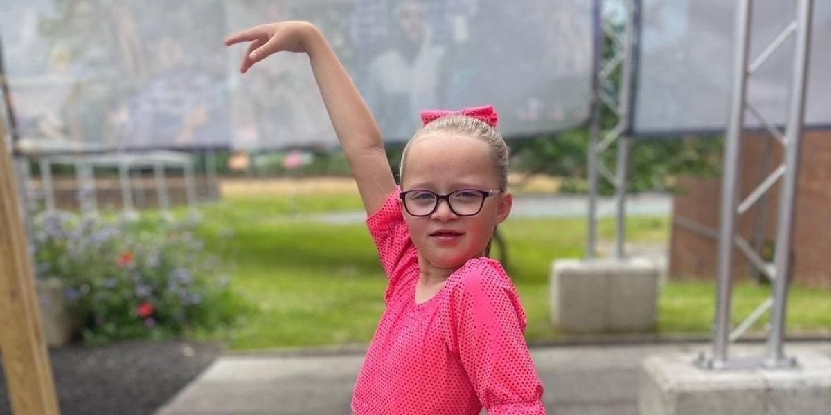 A young girl wearing glasses with one arm up and looking confident.