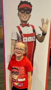 A young girl wearing glasses in a red outfit in front of a big poster.