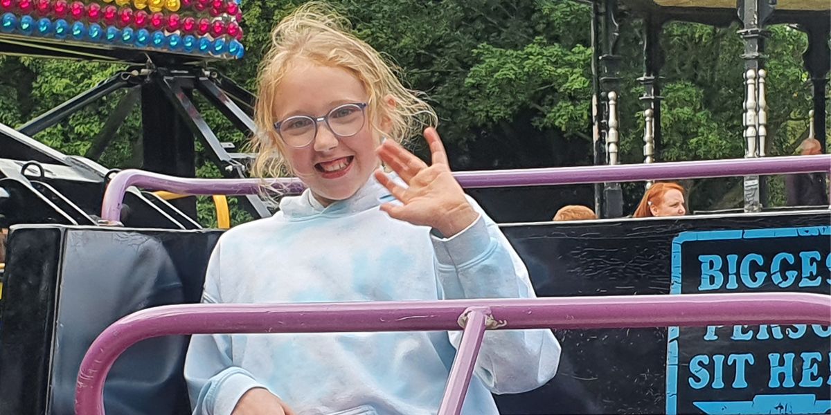 A young girl wearing glasses at a fun fair and waving at the camera.