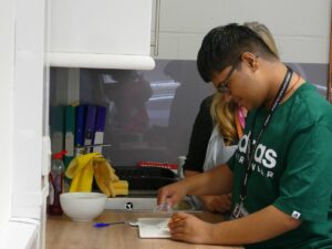 A young man preparing food in a kitchen nect to another person , theyboth have lanyards.