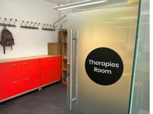A room with a red cabinet and coat hangers. There is an open door with a sign that reads "Therapies room".