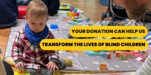 A child say on a playmat surounded by toys and lookind down. Text in black with yellow background says"Your donation can help us transform the lives of blind children."