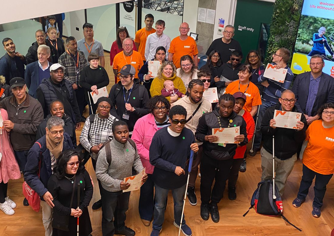 A group of blind and partially sighted children and young people and their families gathered in the RSBC LWLC Atrium holding AQA certificated. They are accompanied with RSBC volunteers, staff and trustees. They are all looking up at the amera.