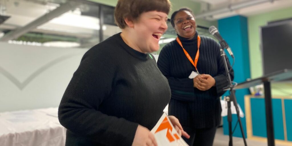 A young woman holding an AQA certificate laughthing with another woman next to her wearing n RSBC lanyard.