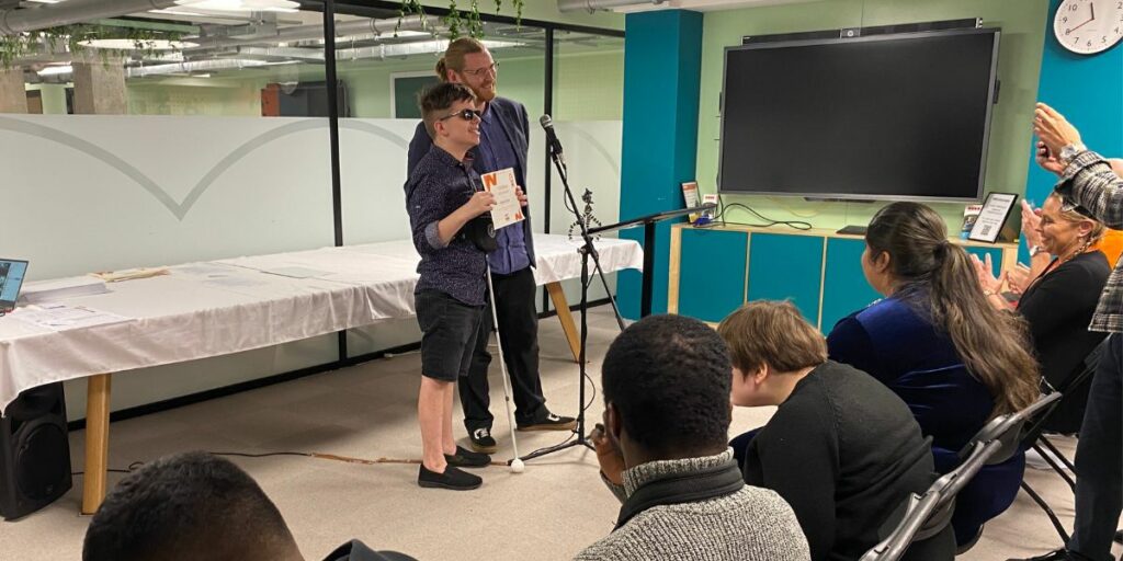 A young person with sight loss wearing glasses and holding his AQA certificate alongside a man standing next to him with a micro in front of him. They are facing an audience sat on chairs.