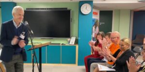 A man with white hair standing up in front of a micro inside a room with people sat on chairs clapping.