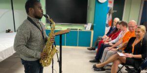 A young person playing saxophone in front of a group of people sat in a room.