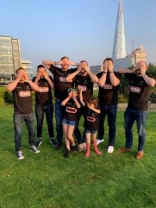 Six men and two little girls standing in a field with their hands over their eyes. They are all wearing black T-shirts.