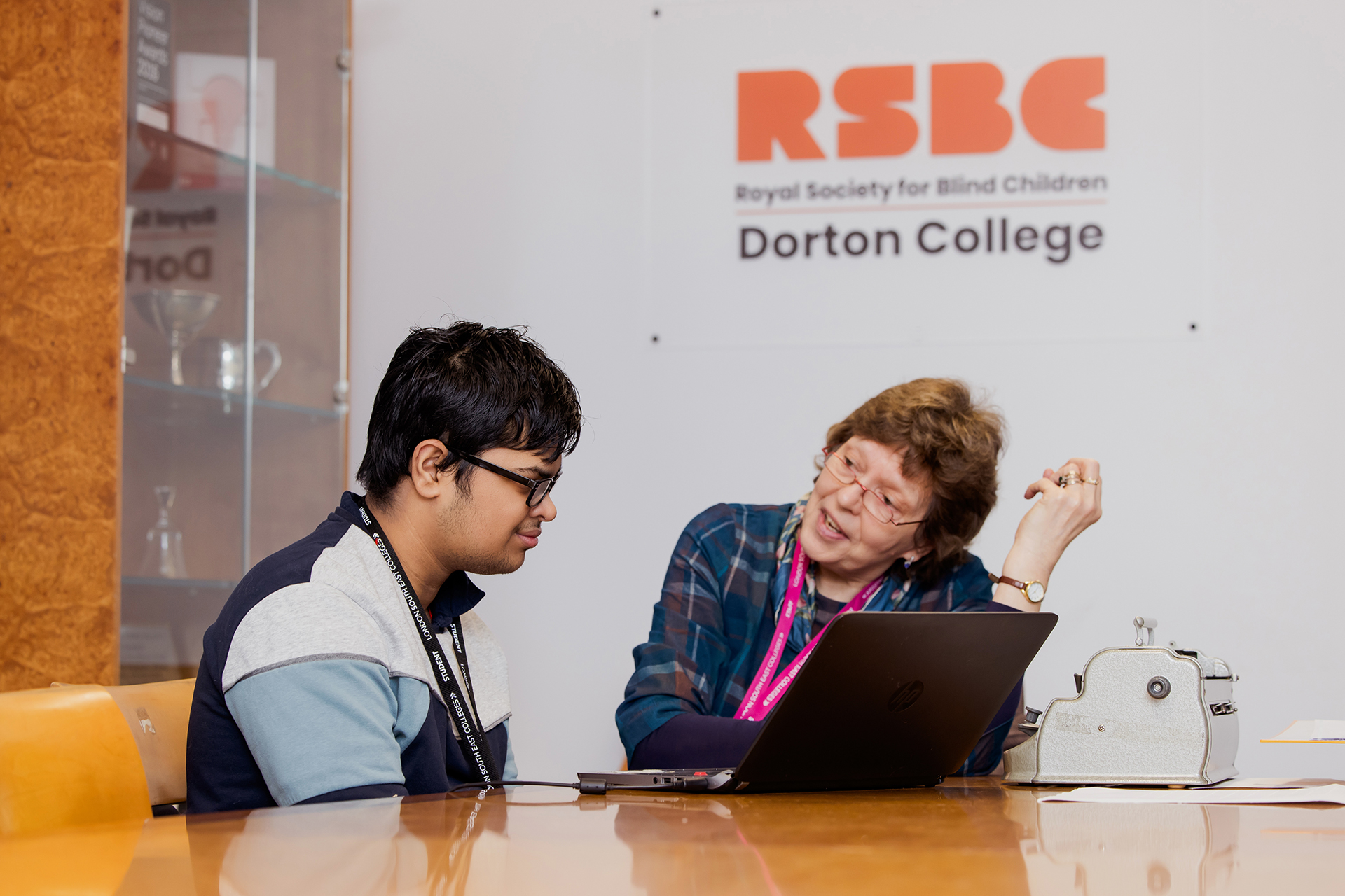 A male student, with glasses, sat down with a female tutor at a desk, listening to her explaining something to him. Behind them on the white wall is an orange RSBC logo with the words Dorton College underneath it.